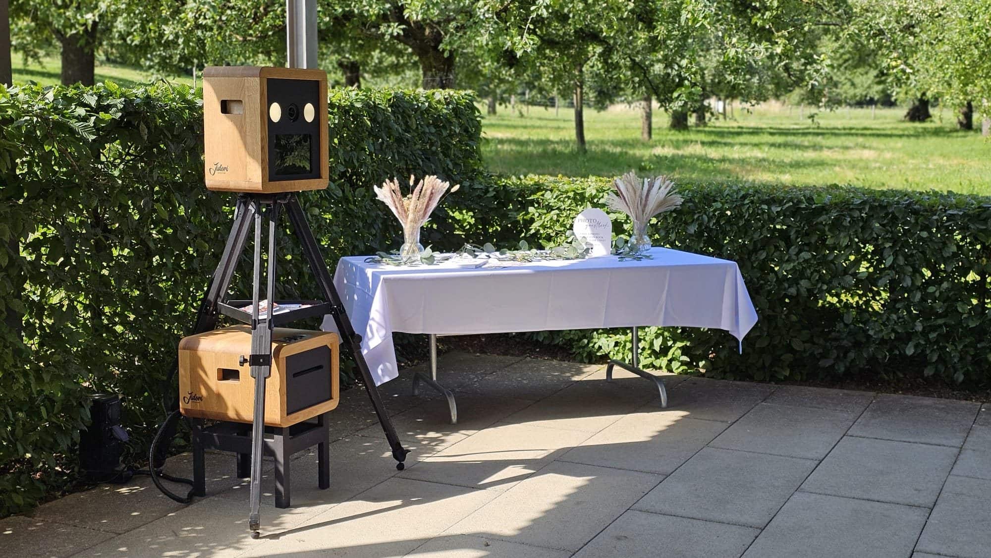 Zwei Jidori-Fotoboxen an einem Gartenfest vor einer Hecke, mit Tisch und Requisiten